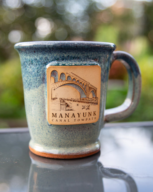 Blue ceramic mug with a bridge logo and 'Manayunk Canal Towpath' text on a reflective surface with blurred green background