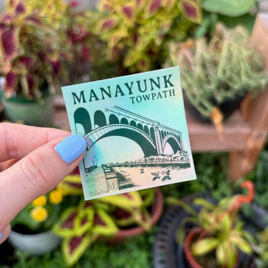 Hand holding a holographic 'Manayunk Towpath' sticker with plants in the background