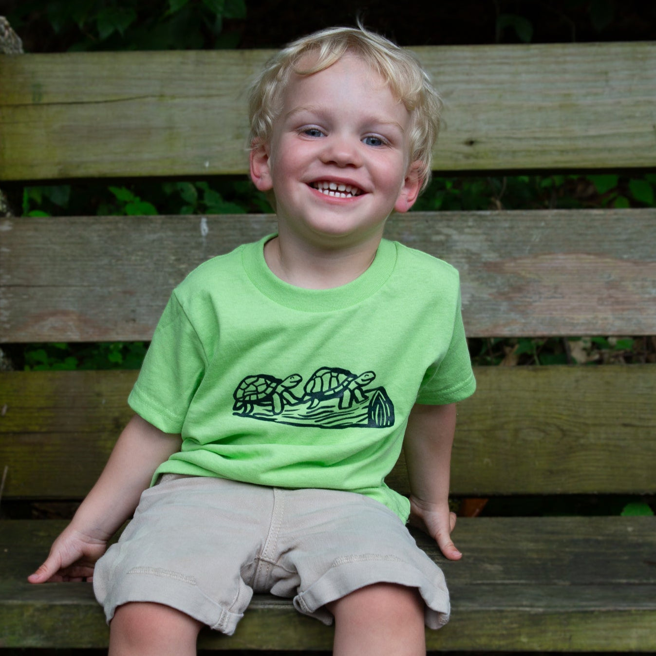 Child wearing a green t-shirt with turtles on a log on it, child is sitting on a wooden bench outdoors.