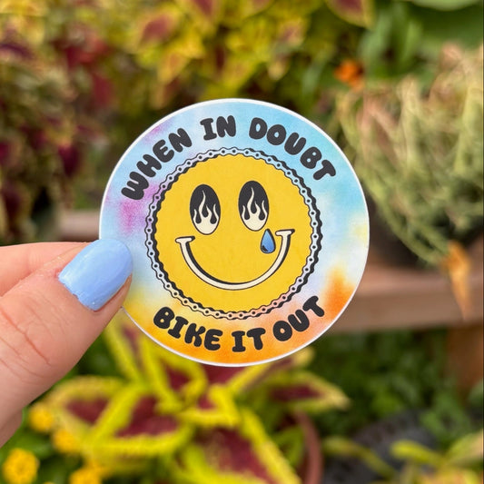 Sticker with smiley face and text 'When in doubt, bike it out' held in front of plants