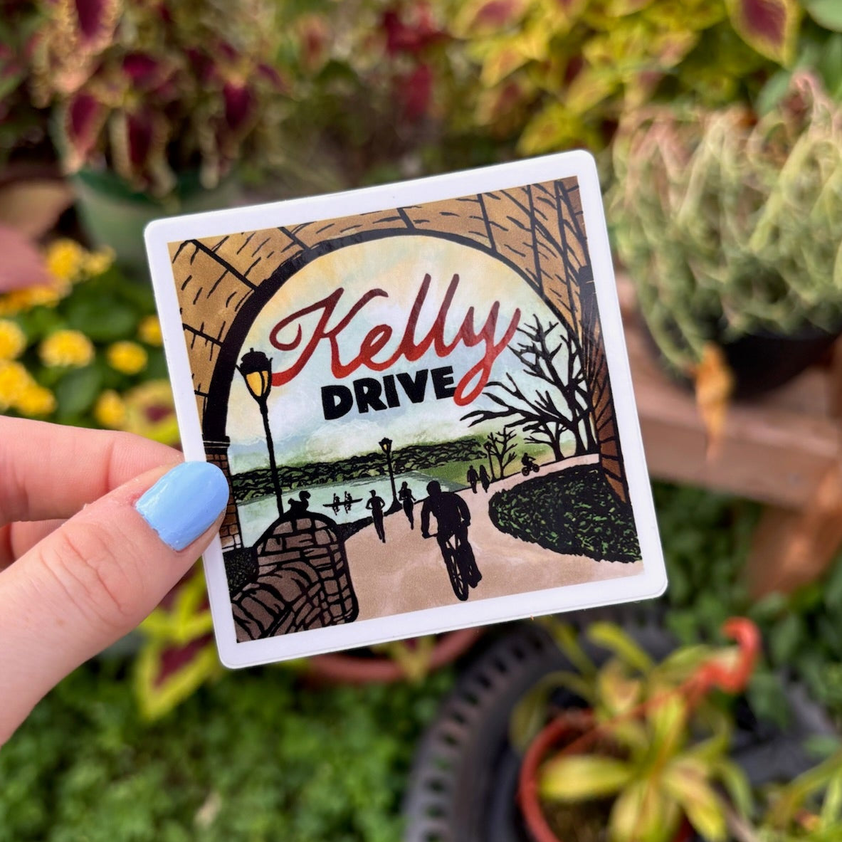 Hand holding a sticker of 'Kelly Drive' design against a garden background
