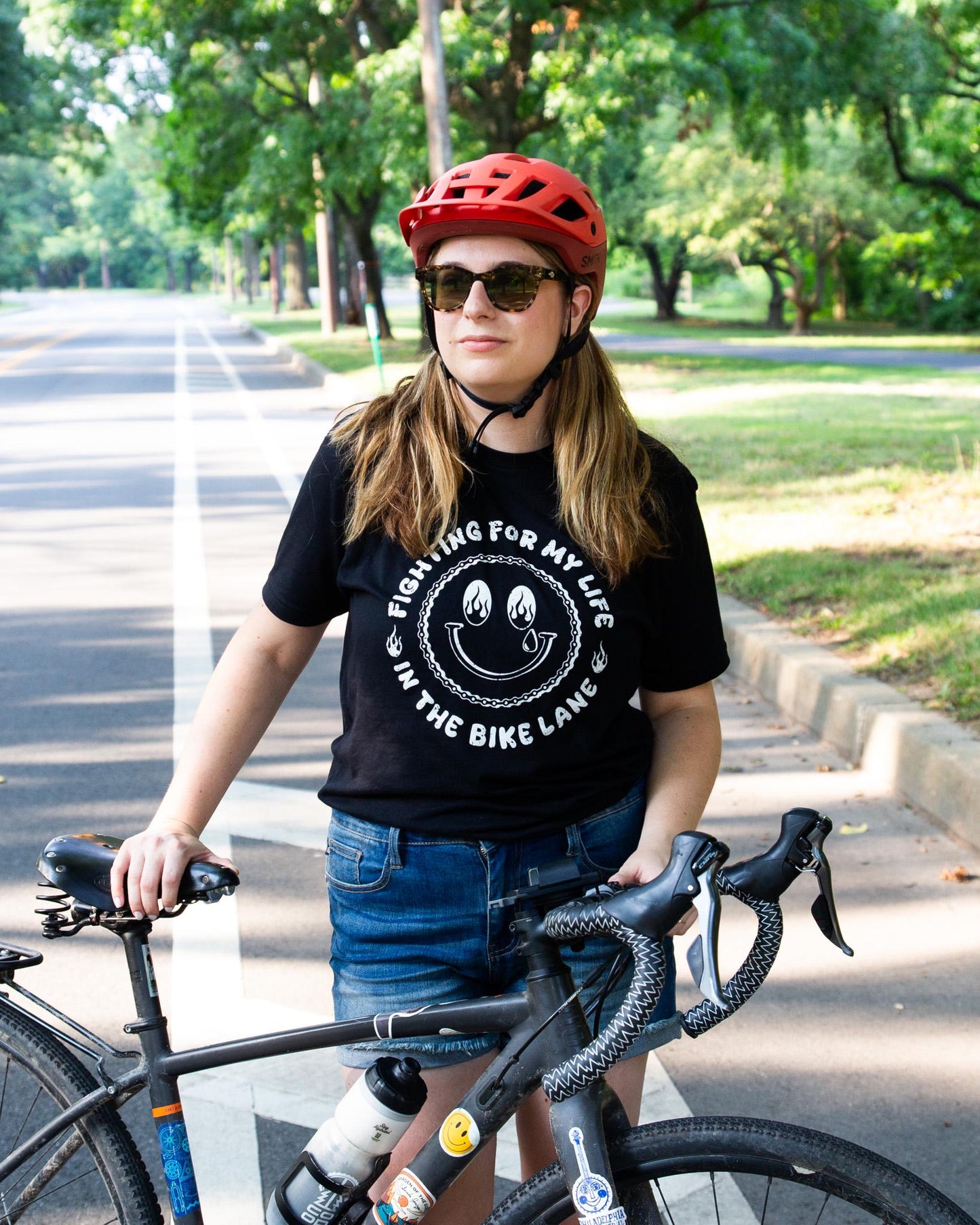 Black Tshirt with a White Illustration of a happy face with Flames in the Eyes that says "Fighting for my life in the bike lane " around the illustration 