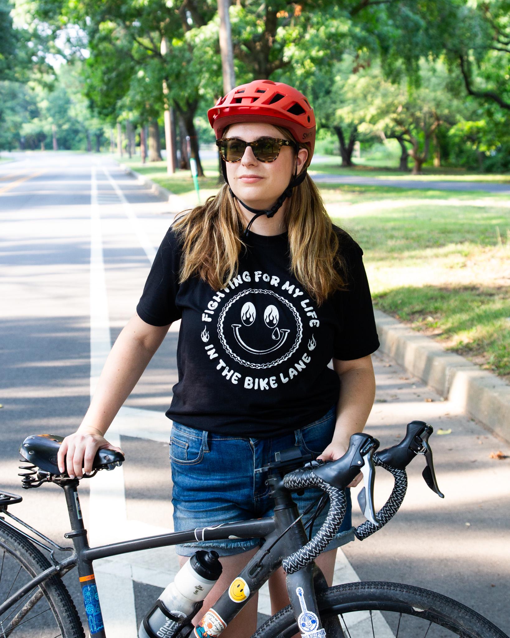 Black Tshirt with a White Illustration of a happy face with Flames in the Eyes that says "Fighting for my life in the bike lane " around the illustration 