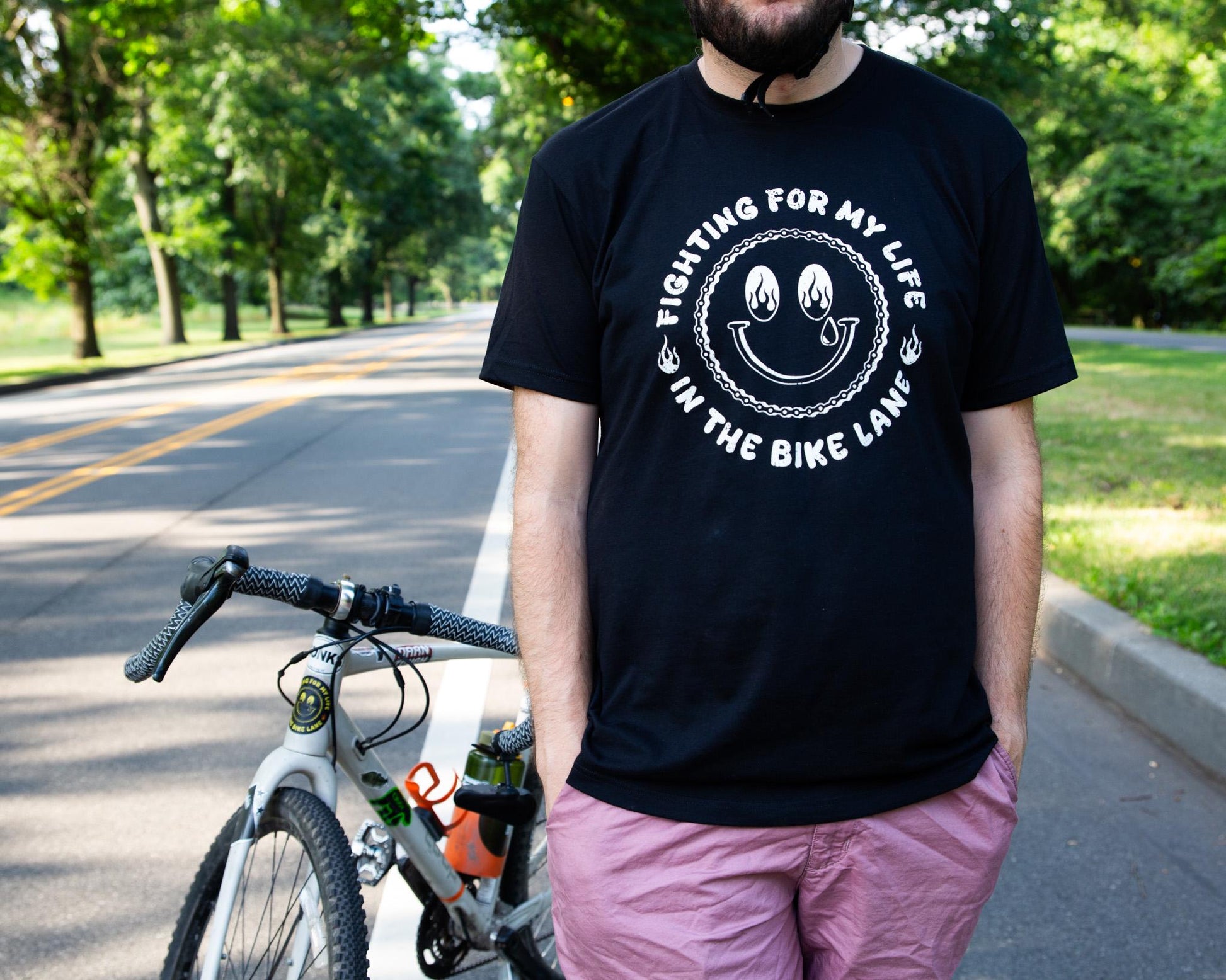 Black Tshirt with a White Illustration of a happy face with Flames in the Eyes that says "Fighting for my life in the bike lane " around the illustration 