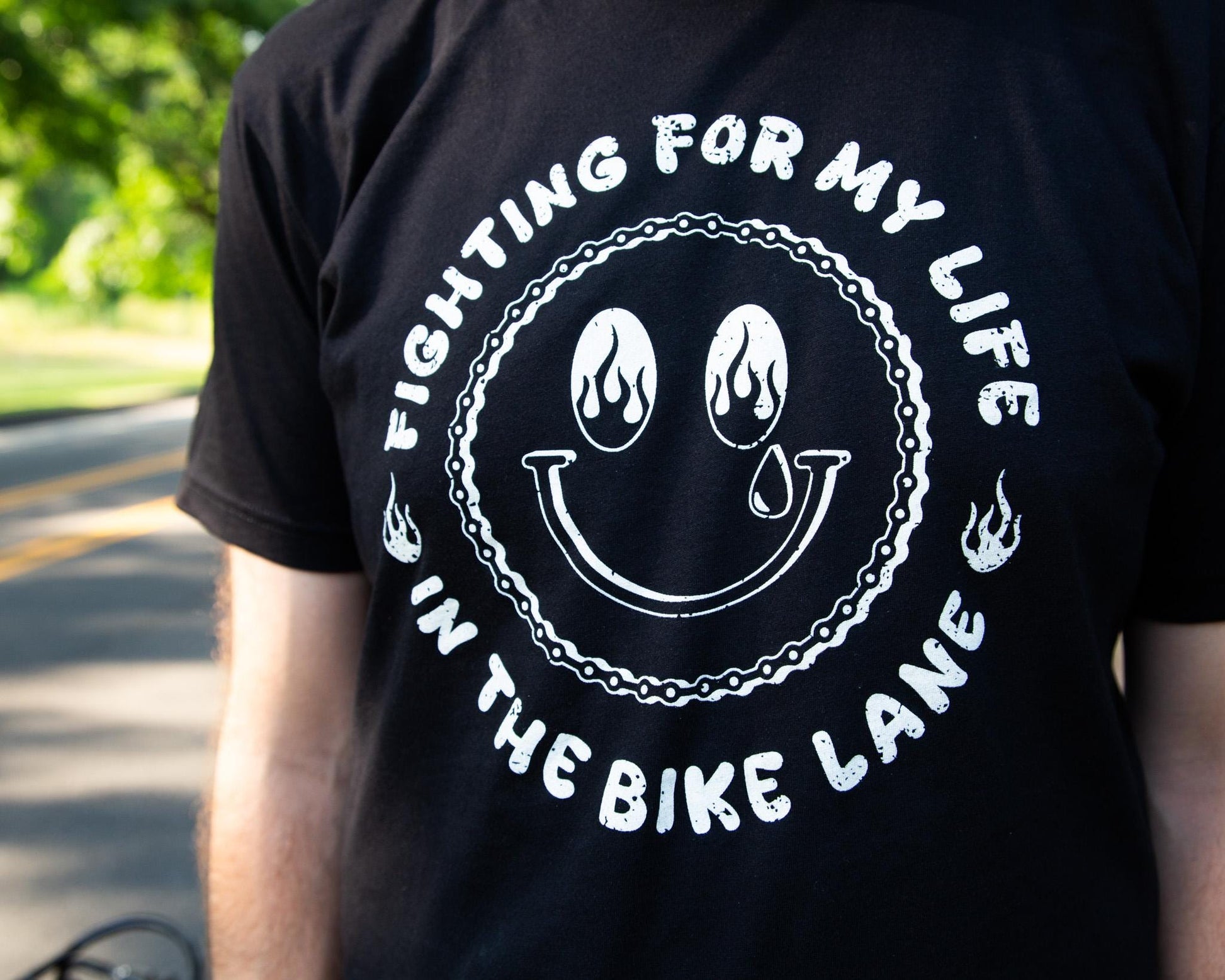 Black Tshirt with a White Illustration of a happy face with Flames in the Eyes that says "Fighting for my life in the bike lane " around the illustration 